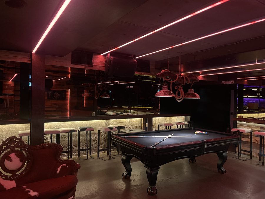 Table de billard sous des néons rouges, queues croisées et banquettes en peau de vache.