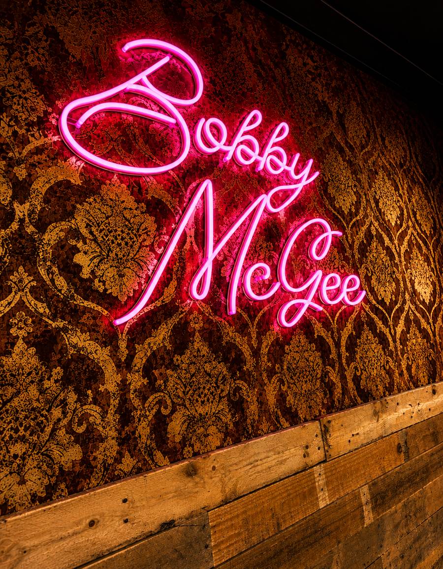Enseigne au néon rose « Bobby McGee » sur un papier peint damassé doré.